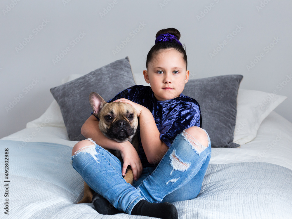Selective focus horizontal portrait of cute blue eyed little girl sitting cross-legged on bed holding her small tan french bulldog Selective focus horizontal portrait of cute blue eyed little girl sitting cross-legged on bed holding her small tan french bulldog