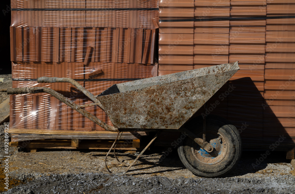 Wheelbarrow at the construction site of a new single-family house building. Unfinished stand-alone home under construction, made of hollow blocks or bricks. Wheelbarrow at the construction site of a new single-family house building. Unfinished stand-alone home under construction, made of hollow blocks or bricks.