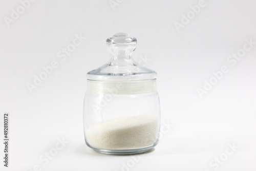 Transparent glass jar with white refined crumbly granulated sugar inside, in the middle on a light background isolated