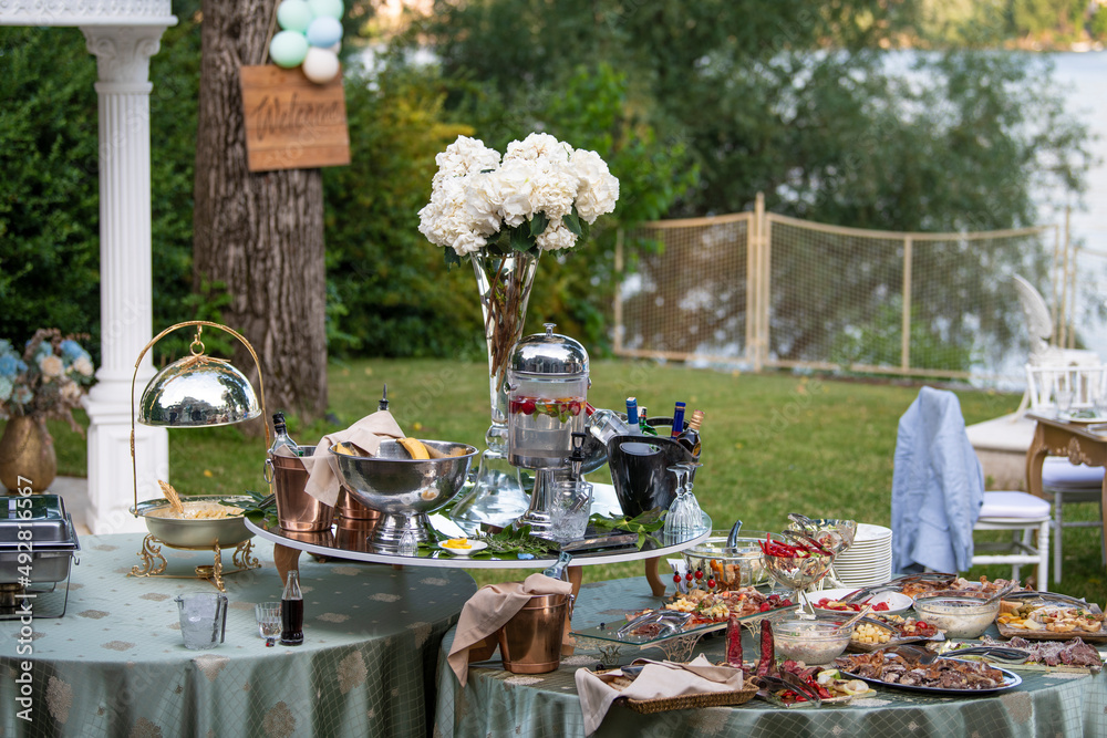Beautiful rich banquet buffet. Table full of different food, snacks,  coctails and drinks, decorated with flowers in rustic style in garden at  party or wedding. Welcome table sign in background. Stock Photo |