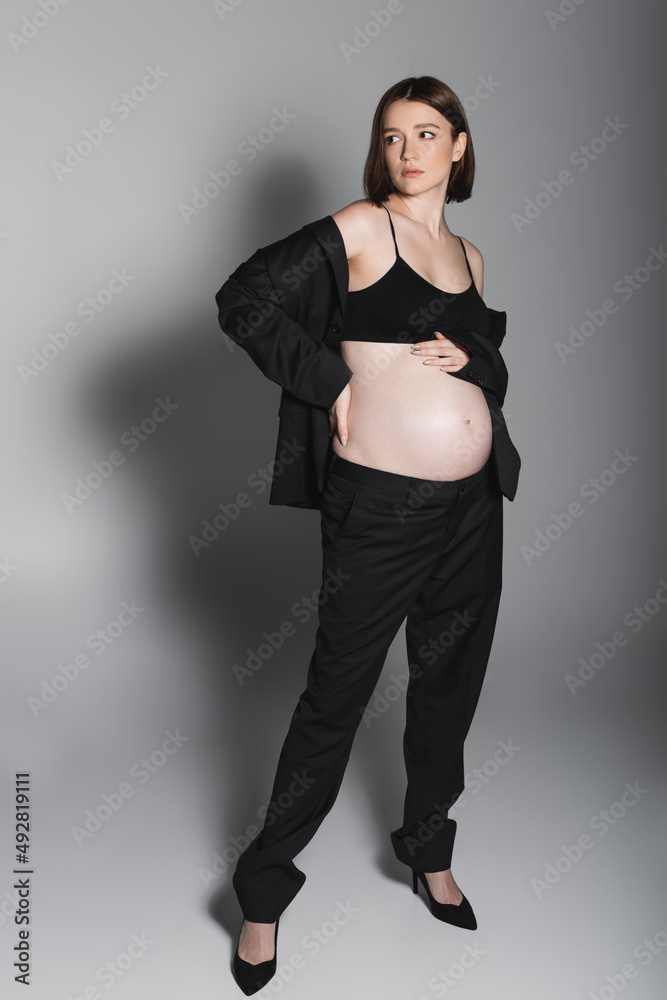 Full length of pregnant woman touching hip and belly on grey background Full length of pregnant woman touching hip and belly on grey background