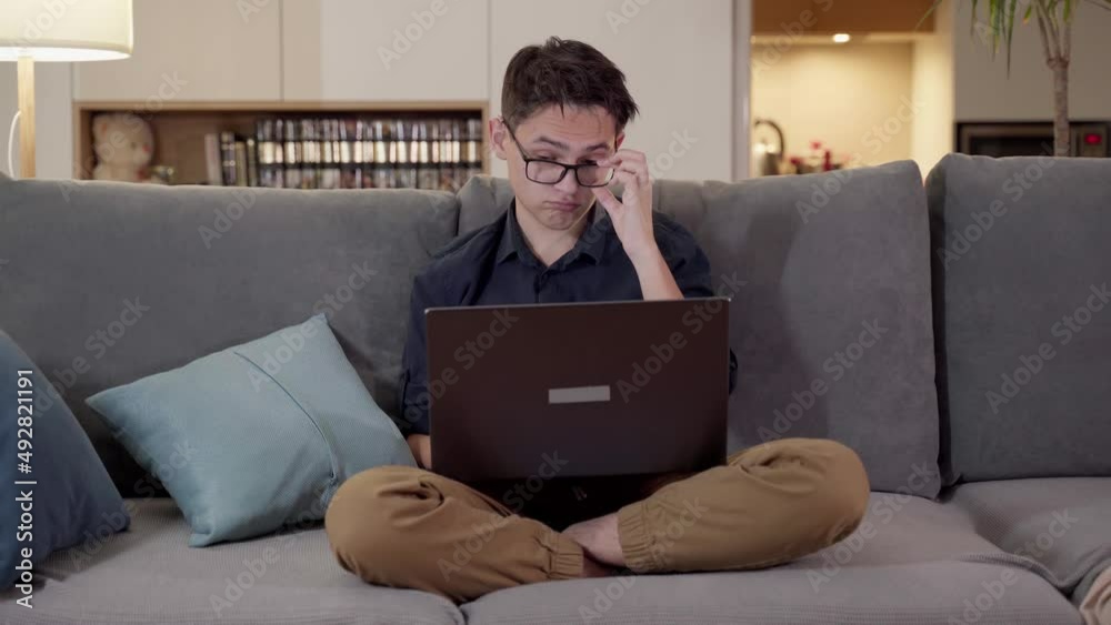 A young guy with glasses sits on a sofa with a laptop. Remote work, online.