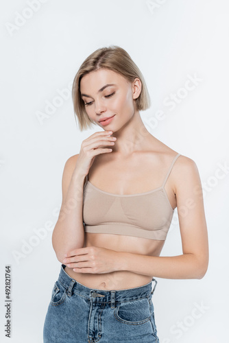 Young blonde woman in top posing and touching chin isolated on grey.