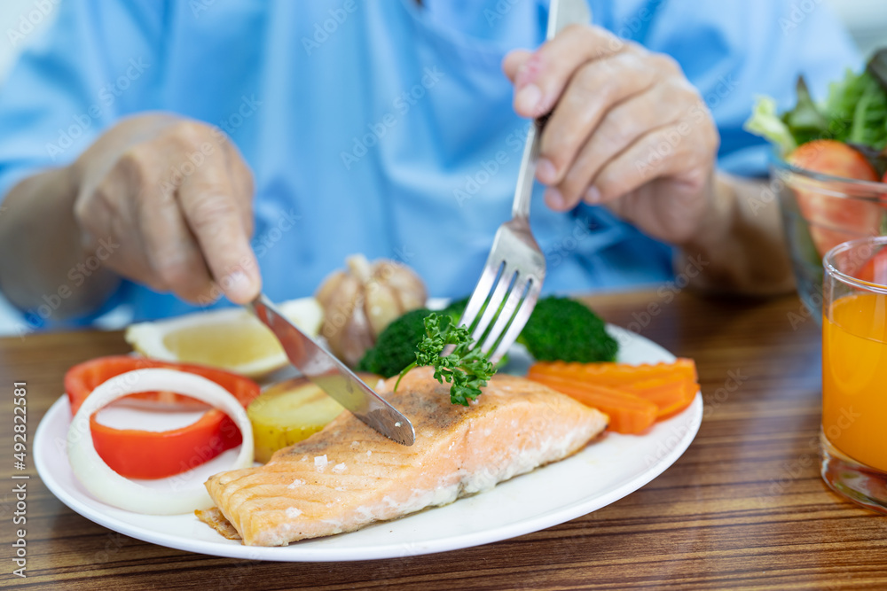 Asian senior or elderly old lady woman patient eating Salmon steak breakfast with vegetable healthy food while sitting and hungry on bed in hospital. Asian senior or elderly old lady woman patient eating Salmon steak breakfast with vegetable healthy food while sitting and hungry on bed in hospital.