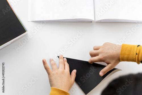 Child doing origami, folding paper on bright white table and shot from above