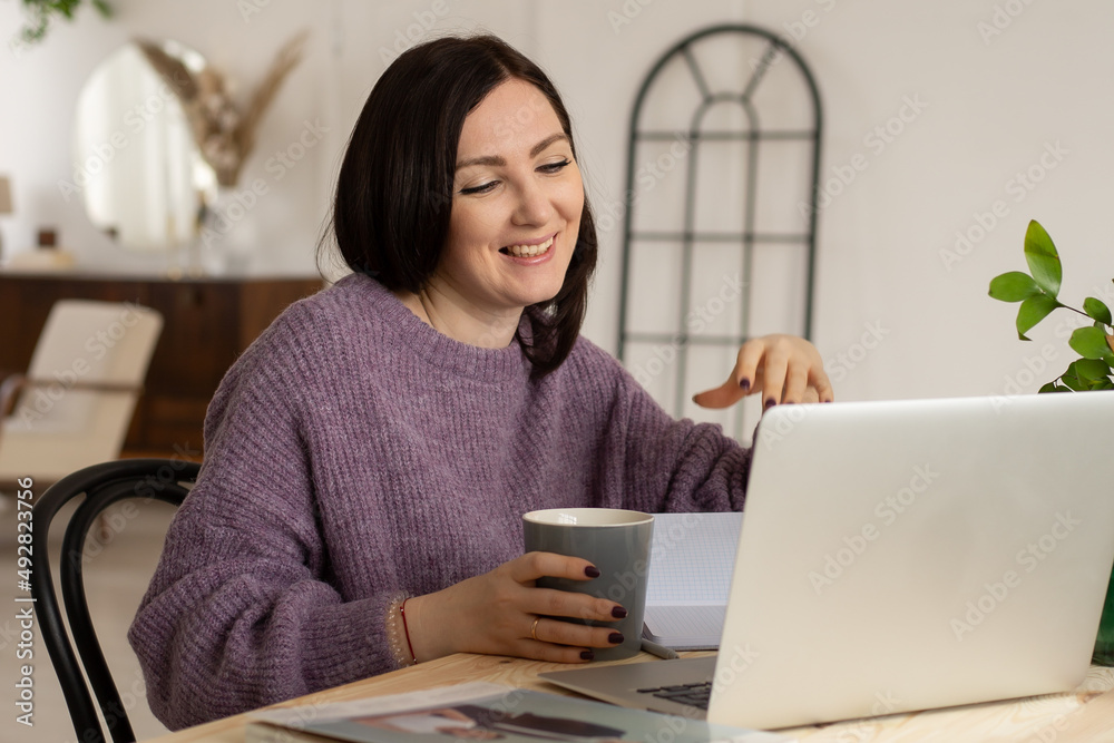 cute brunette girl in a lilac sweater drinks from a gray mug, talks online and smiles, light background, plants on the table cute brunette girl in a lilac sweater drinks from a gray mug, talks online and smiles, light background, plants on the table