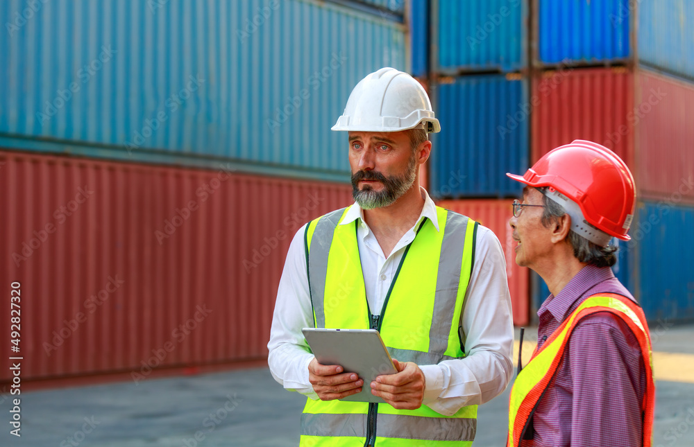 Caucasian and Asian workers teamwork men wear safety clothing. Work clothes with hard hats and use a tablet to inspect containers Caucasian and Asian workers teamwork men wear safety clothing. Work clothes with hard hats and use a tablet to inspect containers