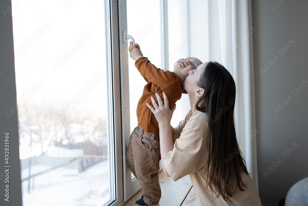 A mother and a small child stand at a large white window and look out into the street. Safety for small children in the house. - obrazy, fototapety, plakaty 