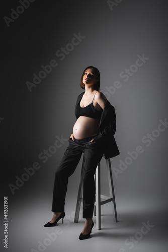 Stylish pregnant woman in heels and jacket sitting on chair on dark grey background