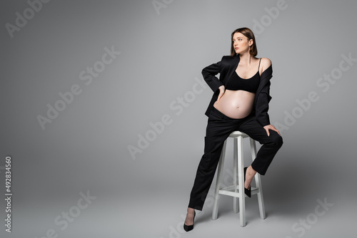 Pregnant woman in black suit posing on chair on grey background