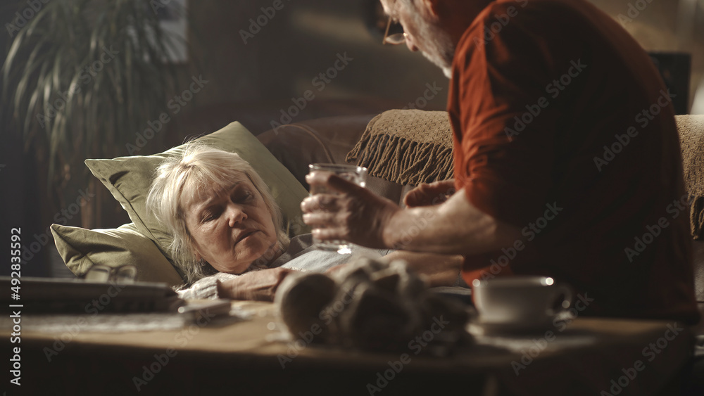 Caring senior husband in glasses giving pill and water to ill wife lying on couch in cozy living room at home Caring senior husband in glasses giving pill and water to ill wife lying on couch in cozy living room at home