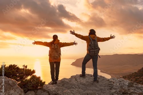 Couple of two young hikers stands with raised arms at mountain top and looks at sea and sunset