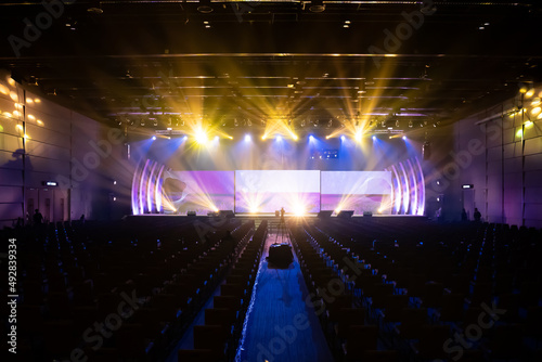 Distant view of the front of the stage with LED screens and spotlights are shining on the stage while testing the lighting system with few staff and many empty chairs arranged for audience in the hall