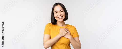 Romantic asian girfriend, holding hands on heart, smiling with care and tenderness, standing in yellow tshirt over white background photo