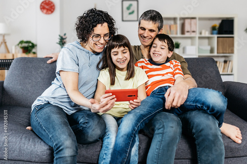 Young beautiful happy family relaxing together while watching funny video or movie on mobile phone while sitting on couch at living room