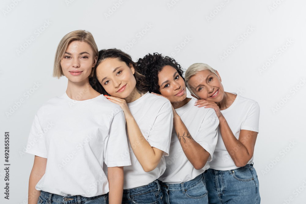 Positive multiethnic women hugging each other and looking at camera isolated on grey. Positive multiethnic women hugging each other and looking at camera isolated on grey.