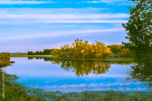 Picturesque view of Midwestern lake with trees in background at sunset in fall; trees and blue sky with light clouds reflect in calm water