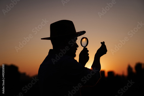 Attractive male silhouette in hat looking with magnifying glass showing small korean heart gesture. Man searching sales or discounts at sunset using loupe with urban view. Investigation concept