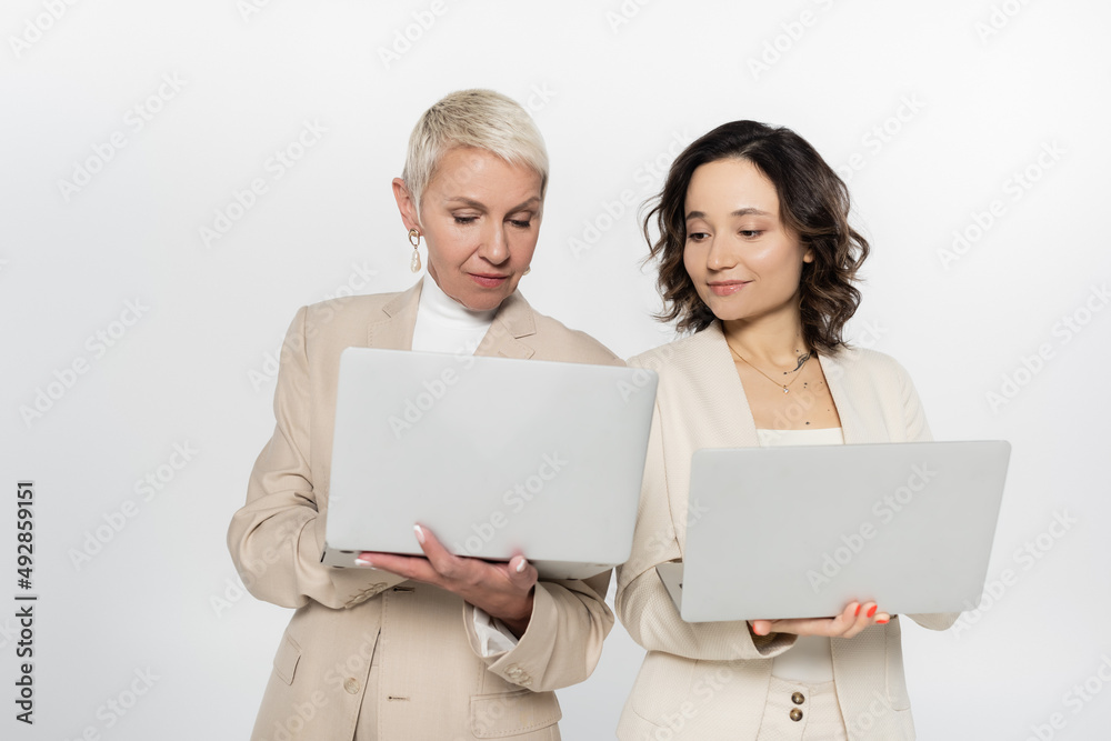 Senior businesswoman holding laptop near colleague isolated on grey. Senior businesswoman holding laptop near colleague isolated on grey.