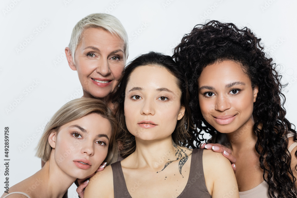 Obraz premium Smiling interracial women hugging tattooed friend isolated on grey, feminism concept.