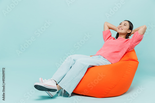 Full body young smiling minded fun woman of Asian ethnicity 20s wear pink sweater sit in bag chair hold hands behind neck isolated on pastel plain light blue color background People lifestyle concept. photo