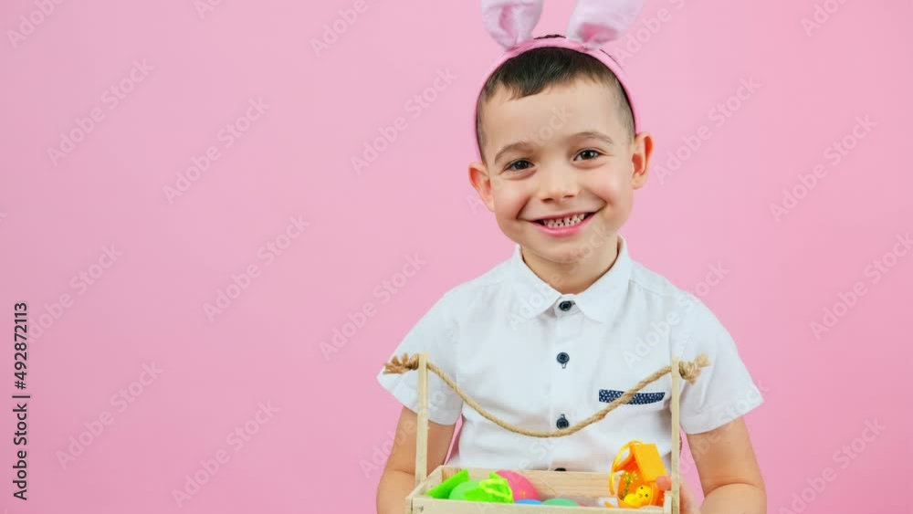 Little cute boy wearing rabbit ears sitting on a chair holding basket with Easter eggs on pink background and copy space. Little cute boy wearing rabbit ears sitting on a chair holding basket with Easter eggs on pink background and copy space.