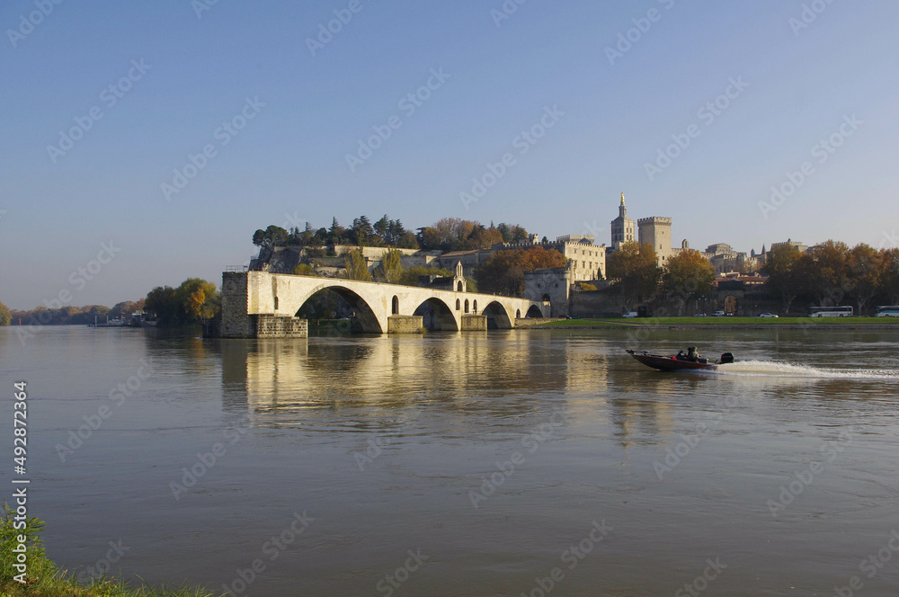 Plan large au lever du jour d'une barque motorisée filant sur le Rhône, au pied du Pont d'Avignon avec vue en arrière plan sur le palais des Papes