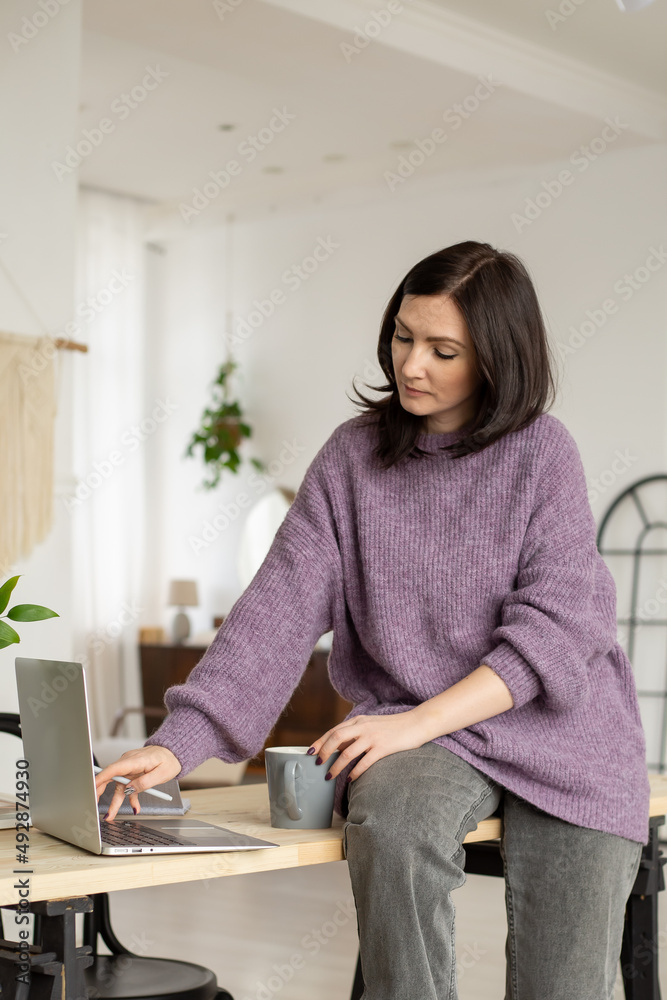 a cute brunette girl in a lilac sweater is sitting on a table with a laptop, looking at the screen and typing a cute brunette girl in a lilac sweater is sitting on a table with a laptop, looking at the screen and typing