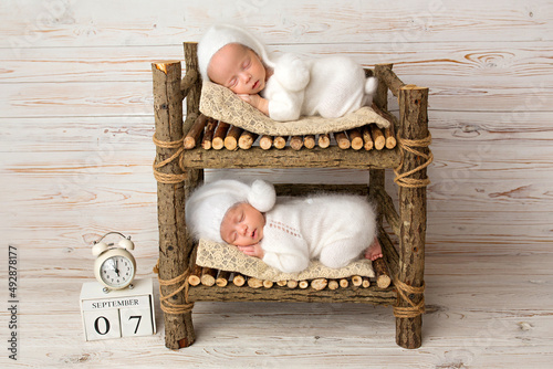 Tiny newborn twin boys in white bodysuits against a light wood background. Newborn twins sleep on a bunk wooden bed. The boys are dressed in white caps. Clock with time of birth. Studio photography.