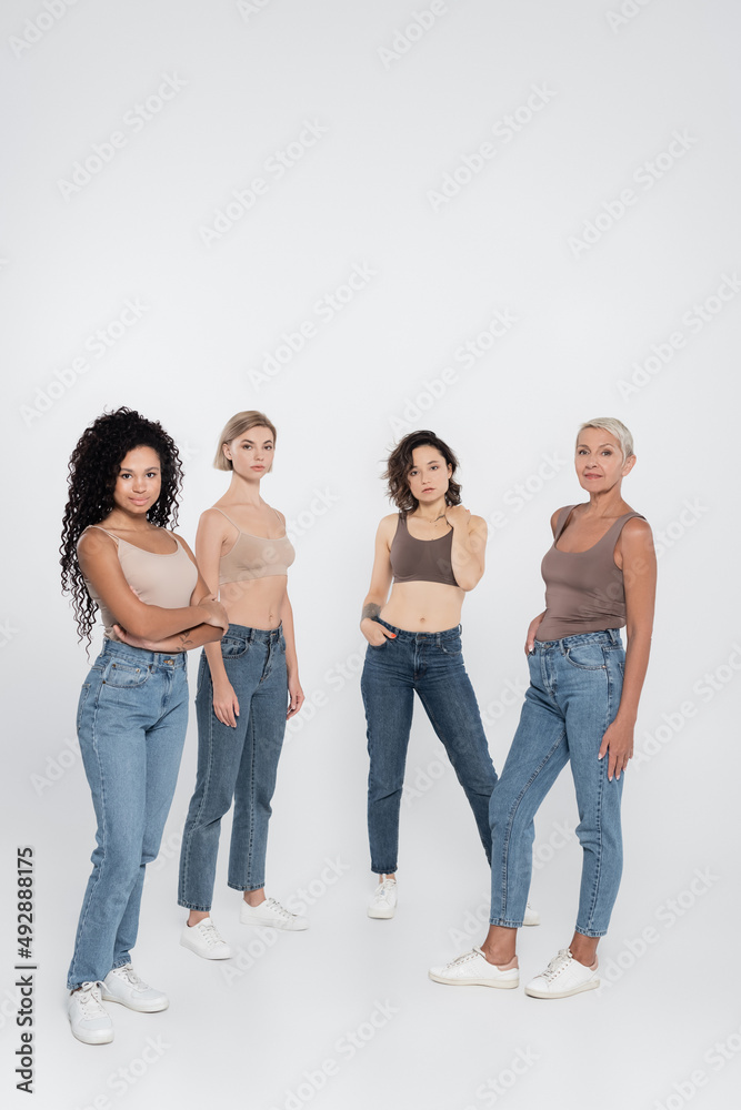 Multiethnic women in jeans posing and looking at camera on grey background.