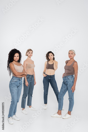 Multiethnic women in jeans posing and looking at camera on grey background.