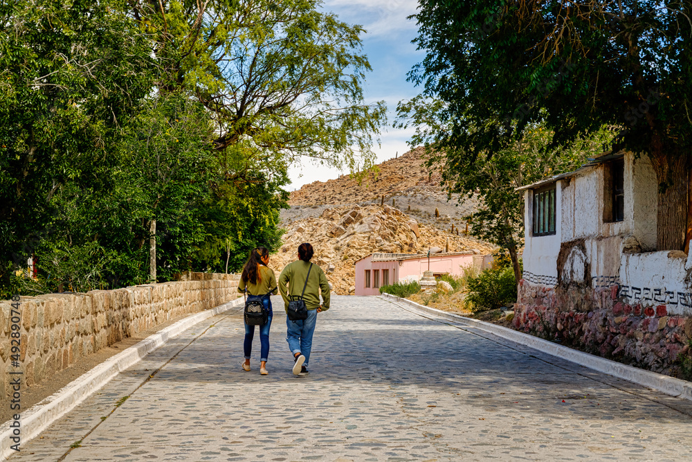 Fototapeta premium Santa Rosa de Tastil, Salta, Argentina - January 6, 2021: Two teenage girls walk down the cobblestone street of the town as they leave school