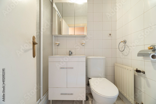 Toilet with white wooden cabinet  cabinet with mirror doors  sink  white tiles and fine aluminum radiator