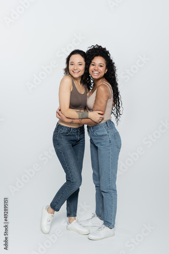 Smiling african american woman hugging tattooed friend on grey background.