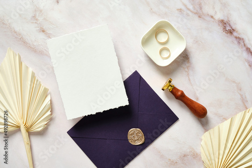 Flat lay wedding stationery on marble table. Blank wedding invitation card mockup, dried flowers, rings, purple envelope, wax seal stamp.