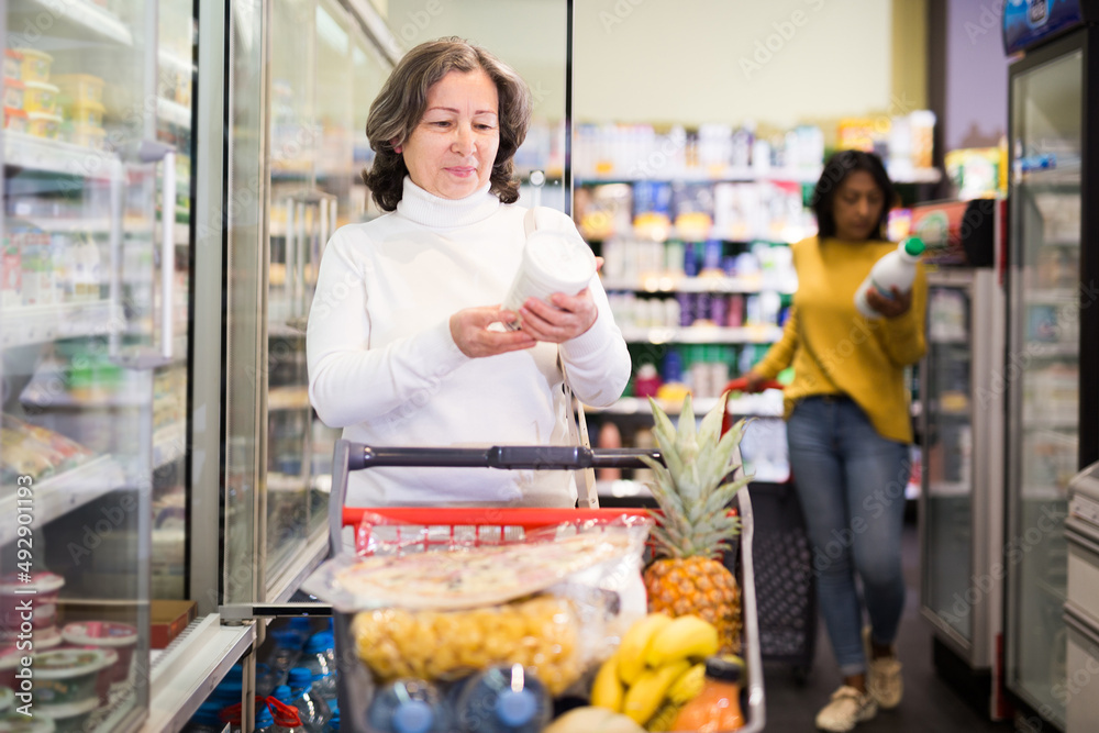 Portrait of interested elderly woman shopping in supermarket, choosing fresh dairy products on refrigerated shelves