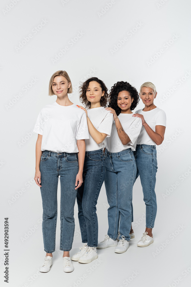 Full length of multiethnic women in jeans and white t-shirts looking at camera on grey background. Full length of multiethnic women in jeans and white t-shirts looking at camera on grey background.
