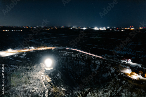 An aerial view of a brightly illuminated cross perched atop a large forested snowy hill during winter at night, in Hamilton, Ontario, Canada.