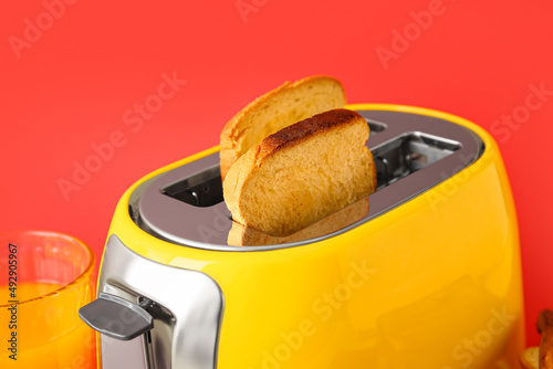 Modern toaster with bread slices and glass of juice on red background, closeup