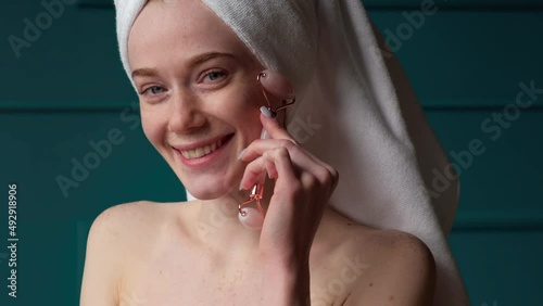 Smiling caucasian woman in bath towel massaging face with jade facial roller to relax muscles. Rejuvenation treatment. Dermatology, cosmetology. Medical photo