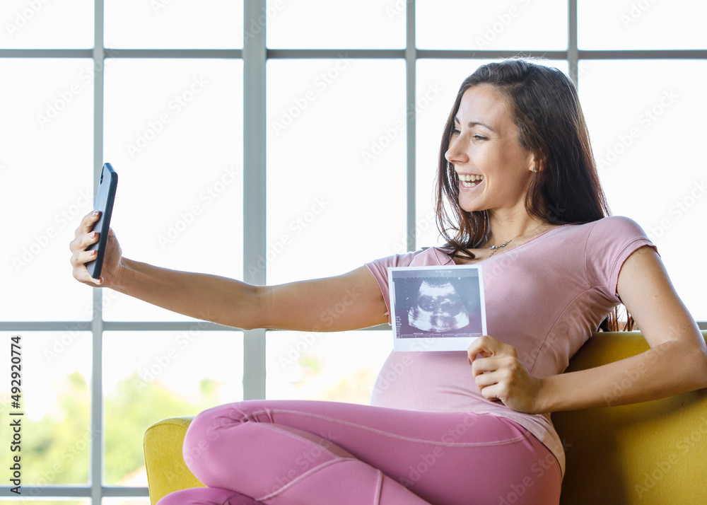 Millennial Caucasian young beautiful happy cheerful healthy big belly tummy pregnancy mother sit on sofa video call conversation via smartphone holding showing ultrasound sonogram Xray film picture