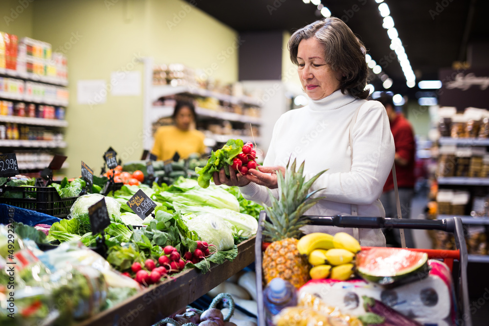 Portrait of positive elderly woman buying fresh organic vegetables in supermarket, choosing red radish