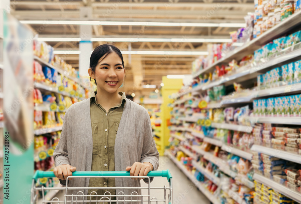 Asian woman shopping from the shelf to the shopping cart in supermarket modern trade hall. Female looking from a grocery store shelf and pick it up to trolley cart.