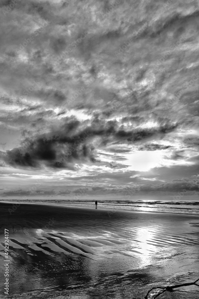 Person in the center of the photo, on the beach sand with a sun rising on the horizon and between clouds. Person in the center of the photo, on the beach sand with a sun rising on the horizon and between clouds.