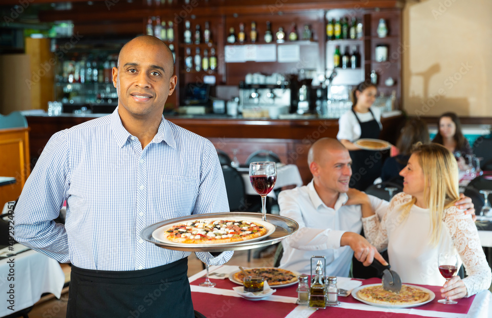 Hospitable latin american waiter standing with serving tray in pizza restaurant hall, welcoming guests