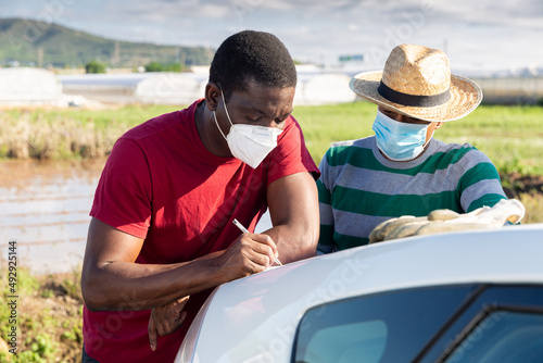 Afro and Latino men farmers wearing disposable face masks signing papers near car on farm