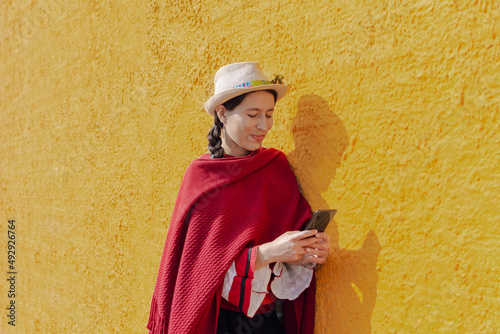 Latin peasant woman in typical costume checking her mobile phone with a yellow rustic wall
