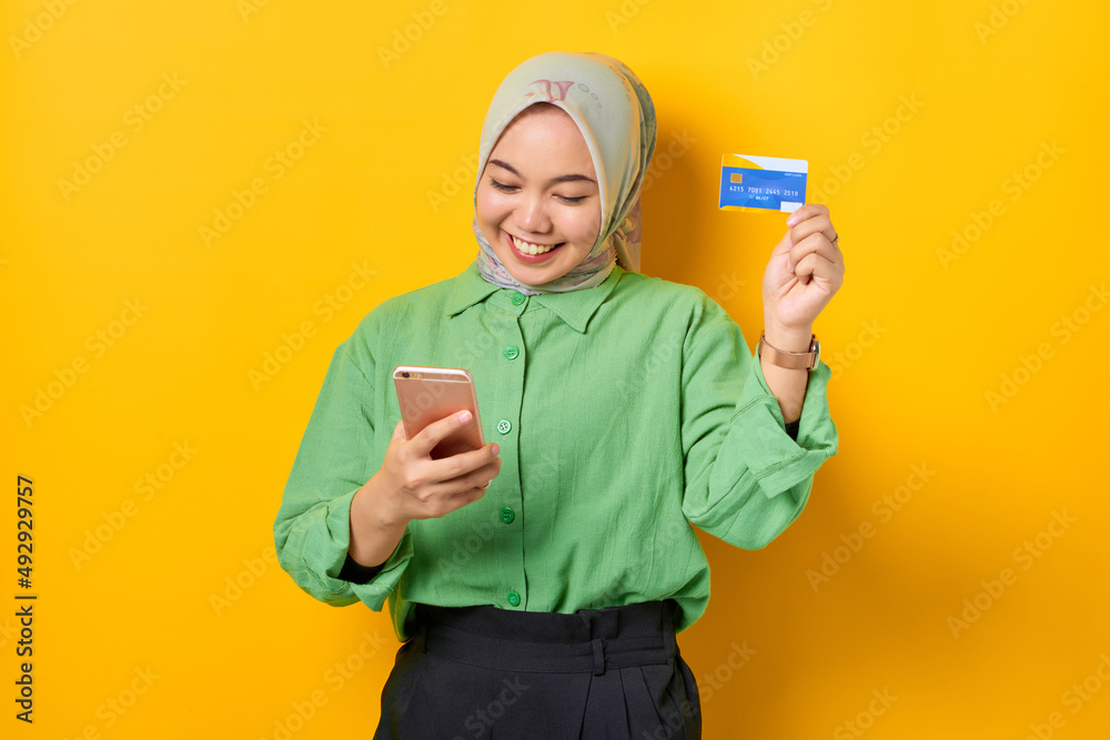 Cheerful young Asian woman in a green shirt holding mobile phone and credit card, rejoicing luck on yellow background