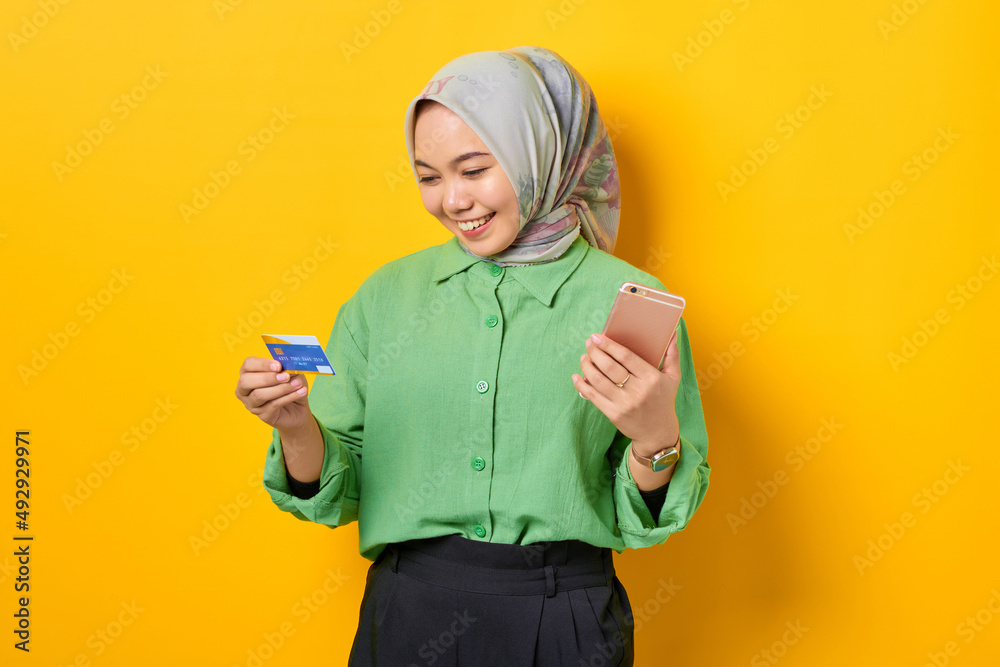 Cheerful young Asian woman in a green shirt holding mobile phone and credit card, rejoicing luck on yellow background