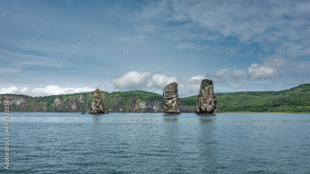 High picturesque cliffs with steep slopes rise above the Pacific Ocean. The green hilly coast of Kamchatka against the background of blue sky and clouds. Rocks Three Brothers